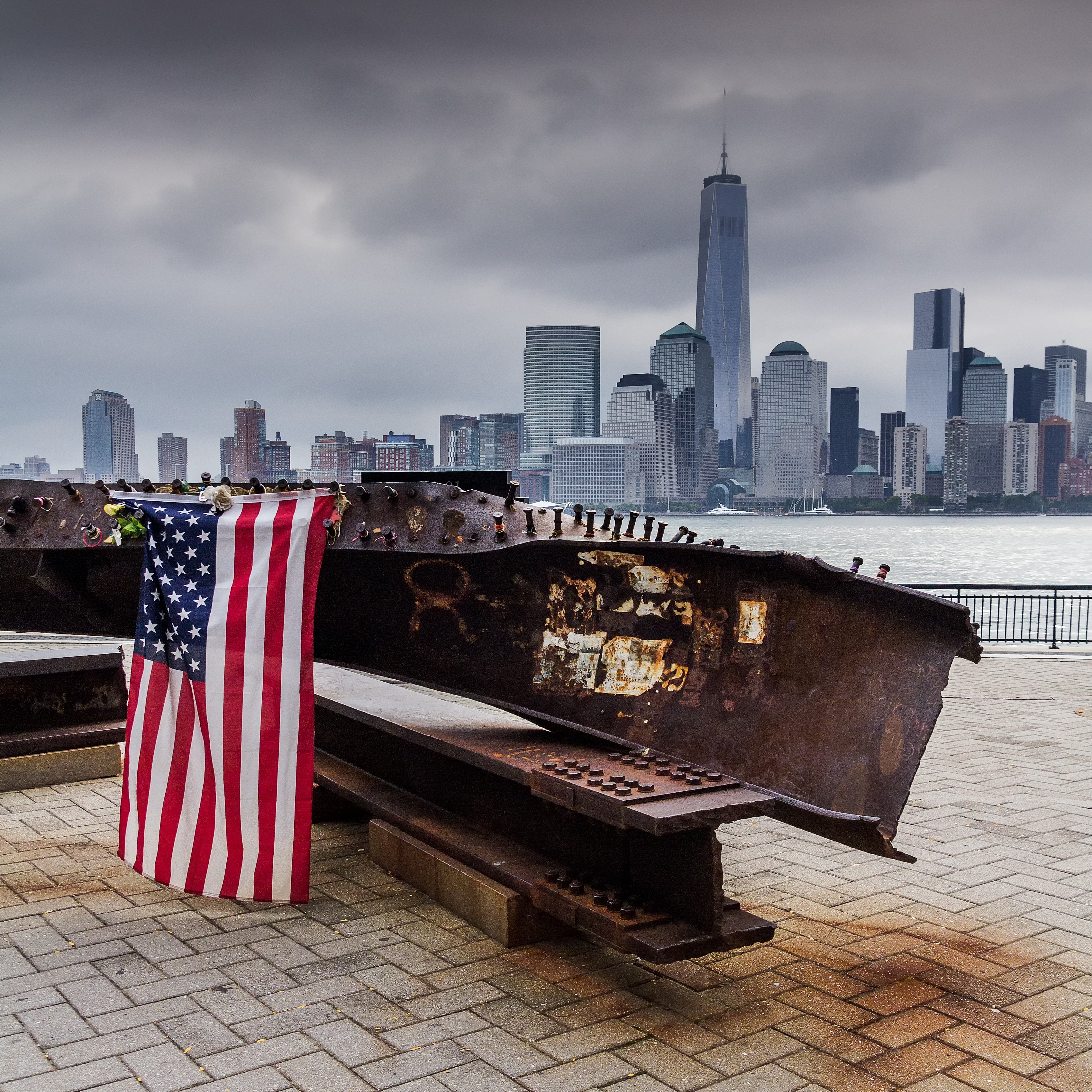 Una trave d'acciaio proveniente da una delle torri del World Trade Center cadute, dall'altra parte del fiume Hudson da Manhattan. Una bandiera americana è drappeggiata sulla trave con la Freedom Tower sullo sfondo.